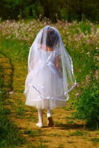 Young girl wearing a white dress and veil walking down a dirt path through a field of wildflowers, dressed for her First Communion.
