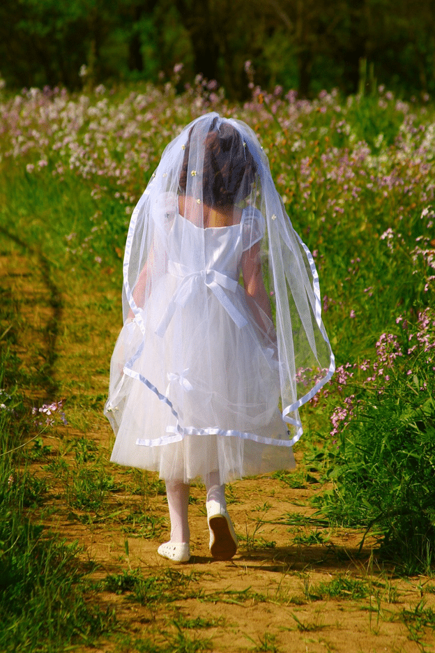 Young girl wearing a white dress and veil walking down a dirt path through a field of wildflowers, dressed for her First Communion.