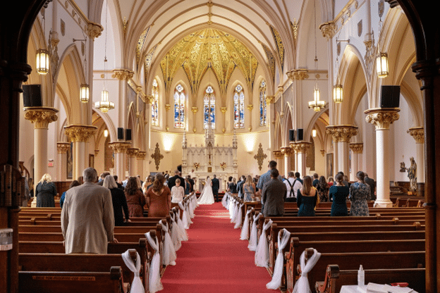 Congregation gathered inside an ornate Catholic church with stained glass windows and gold-accented arches, witnessing a religious ceremony at the altar.