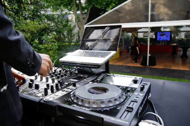 Close-up of a DJ’s hands operating a sound mixer and laptop at an outdoor event under a clear tent, with guests mingling in the background.