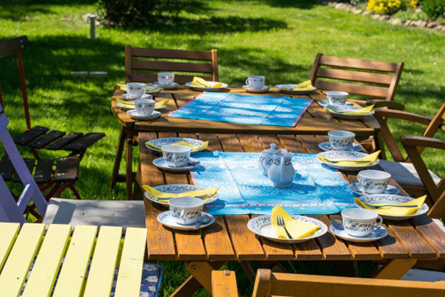 Wooden patio table set for a garden tea party with china teacups, saucers, and yellow napkins, arranged on a sunny lawn with green grass.