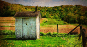 Vintage wooden outhouse with a crescent moon cutout on the door, set in a grassy rural landscape with hills and trees in the background.
