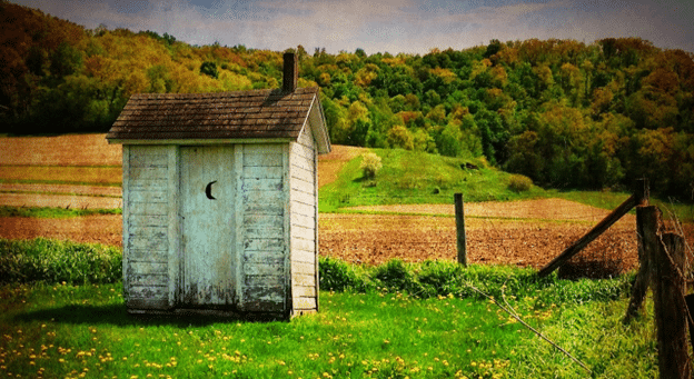 Vintage wooden outhouse with a crescent moon cutout on the door, set in a grassy rural landscape with hills and trees in the background.