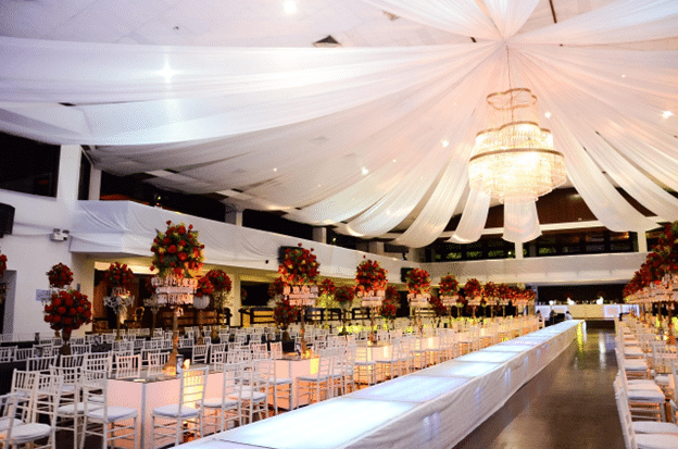 Elegant indoor event venue with long banquet tables, white draped ceiling, crystal chandelier, and tall floral centerpieces arranged for a formal celebration or graduation party.