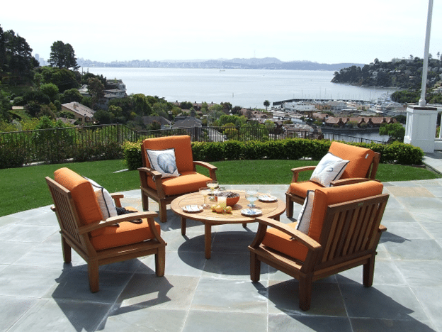 Stylish outdoor patio setup with orange-cushioned wooden chairs arranged around a round table, overlooking a scenic waterfront and hillside community.