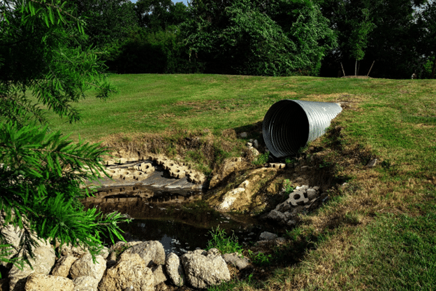 Take the load off: Large metal drainage pipe outlet discharging into a shallow, rocky basin on a grassy slope, surrounded by erosion control blocks and bordered by trees in the background.