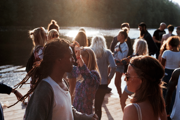 Group of young adults socializing and dancing outdoors on a dock near a lake during sunset, with trees in the background and warm natural light highlighting the scene.