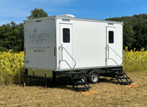 White 5 Unit Bonavita Site Solutions restroom trailer parked in a sunny field beside blooming sunflowers, with dual entry stairs and trees in the background.