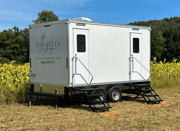 White 5 Unit Bonavita Site Solutions restroom trailer parked in a sunny field beside blooming sunflowers, with dual entry stairs and trees in the background.