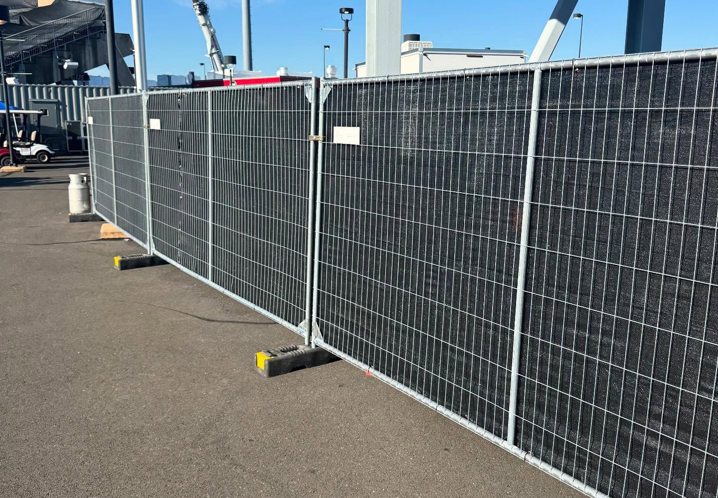 Temporary panel fencing with black privacy mesh and weighted bases at an event venue.