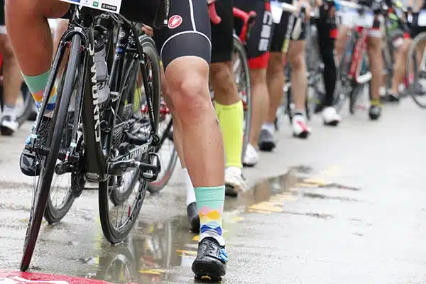 Cyclists lined up at a rainy start line—close-up of bikes and colorful socks on wet pavement at a bike race staging area.