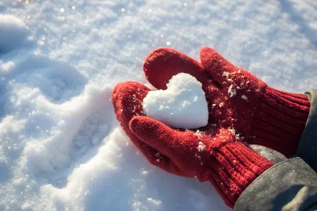 Red mittens holding a heart-shaped snowball on fresh snow.