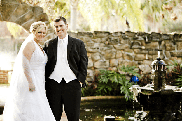 Bride and groom posing beside a garden fountain and stone wall.
