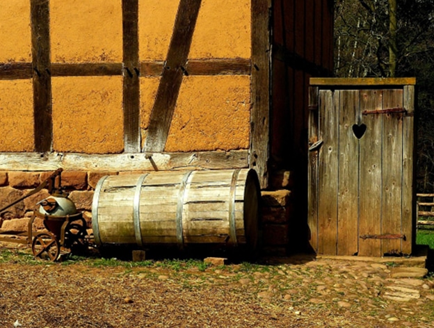 Old wooden outhouse with heart-cutout door beside a rustic barn and barrel.