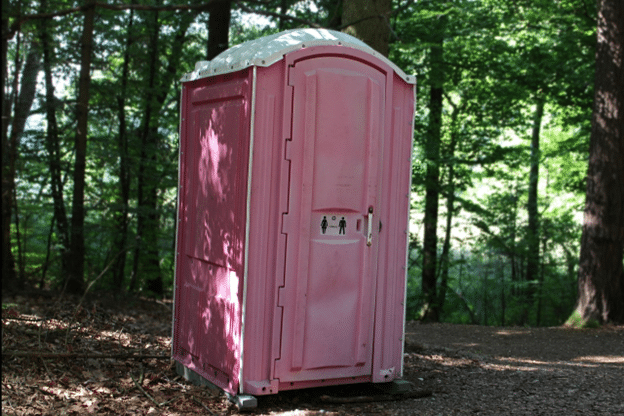 Pink portable restroom set on a forest path in dappled light.