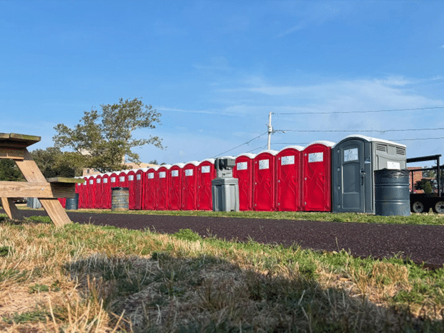 Long row of red portable restrooms with a gray ADA unit and handwash station along a park path.